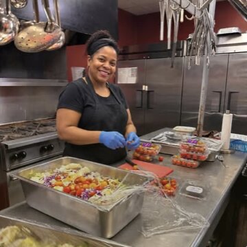 A cook in gloves smiling as she prepares trays of food in the Hosea House kitchen