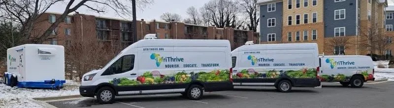 Three AfriThrive delivery vans and a refrigerated trailer parked outside an apartment complex