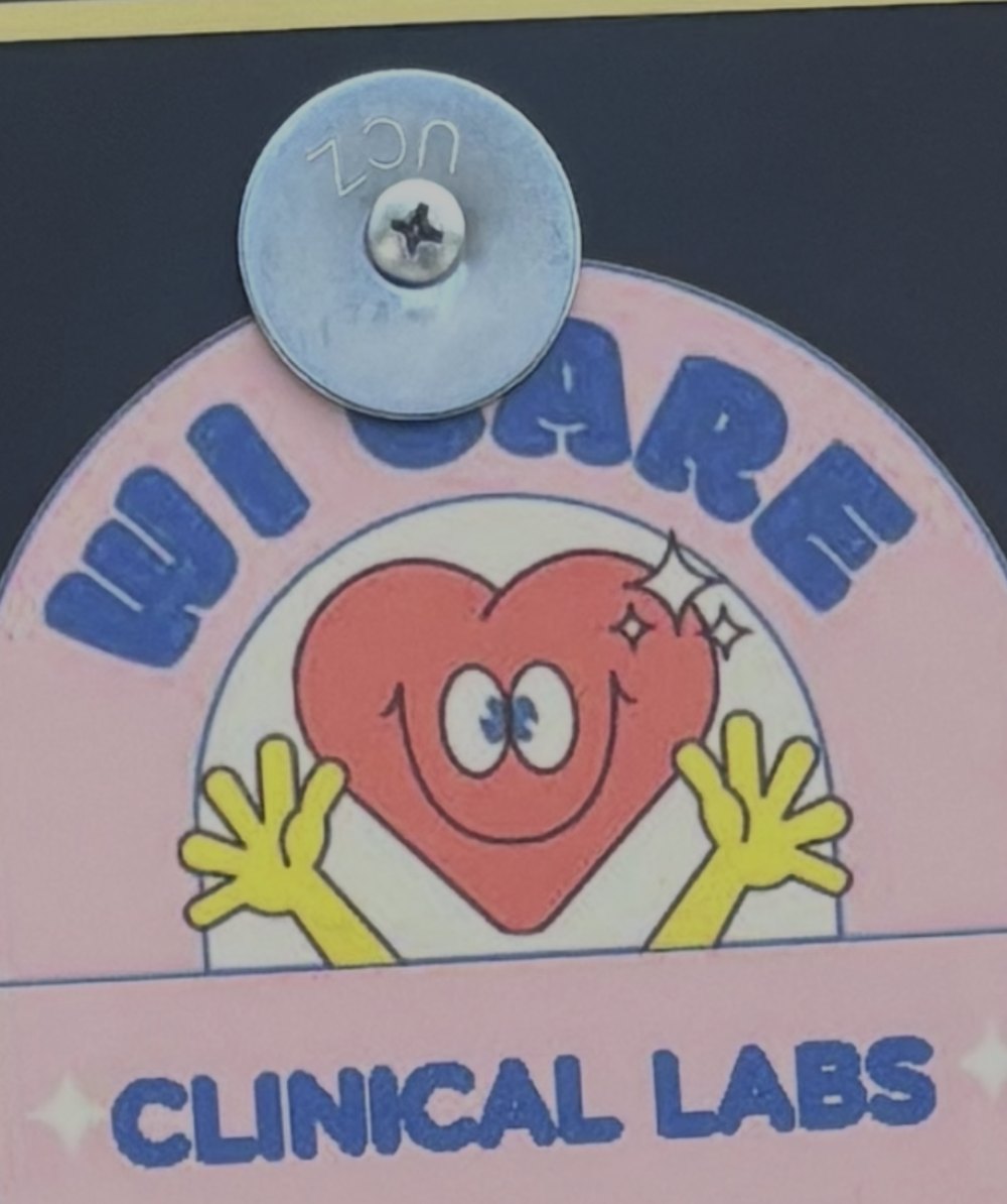 A Wi Care Clinical Labs sign showing a smiling cartoon heart with raised hands on a pink arch