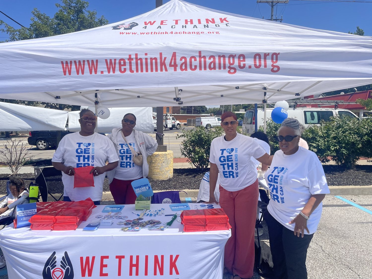 Four volunteers in Together We Can End HIV shirts at a We Think 4 A Change outreach tent with information and giveaways on the table